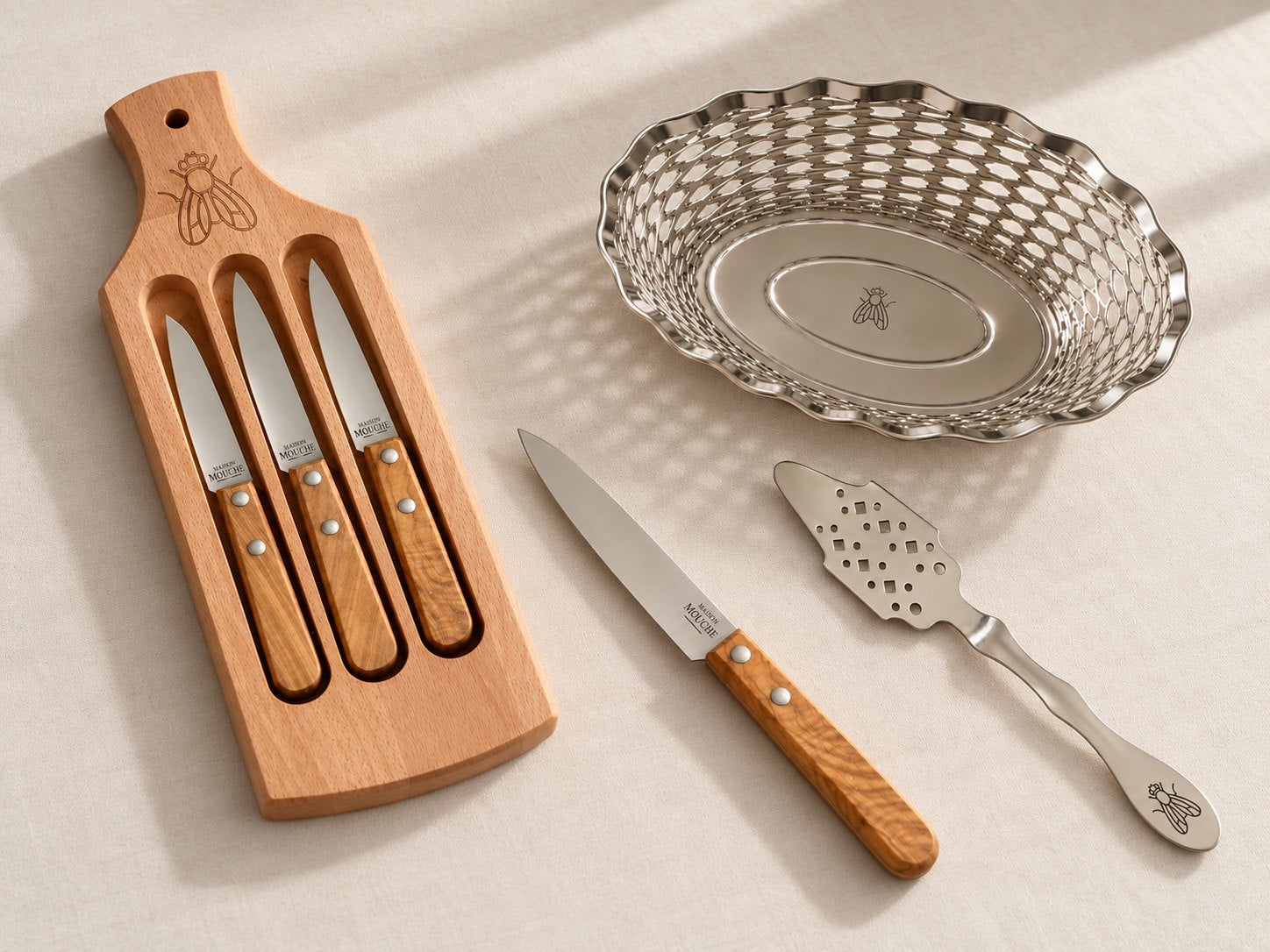 La table Maison Mouche : planche et 3 couteaux de Thiers, corbeille à pain en métal ajouré, couteau de table et cuillère à absinthe